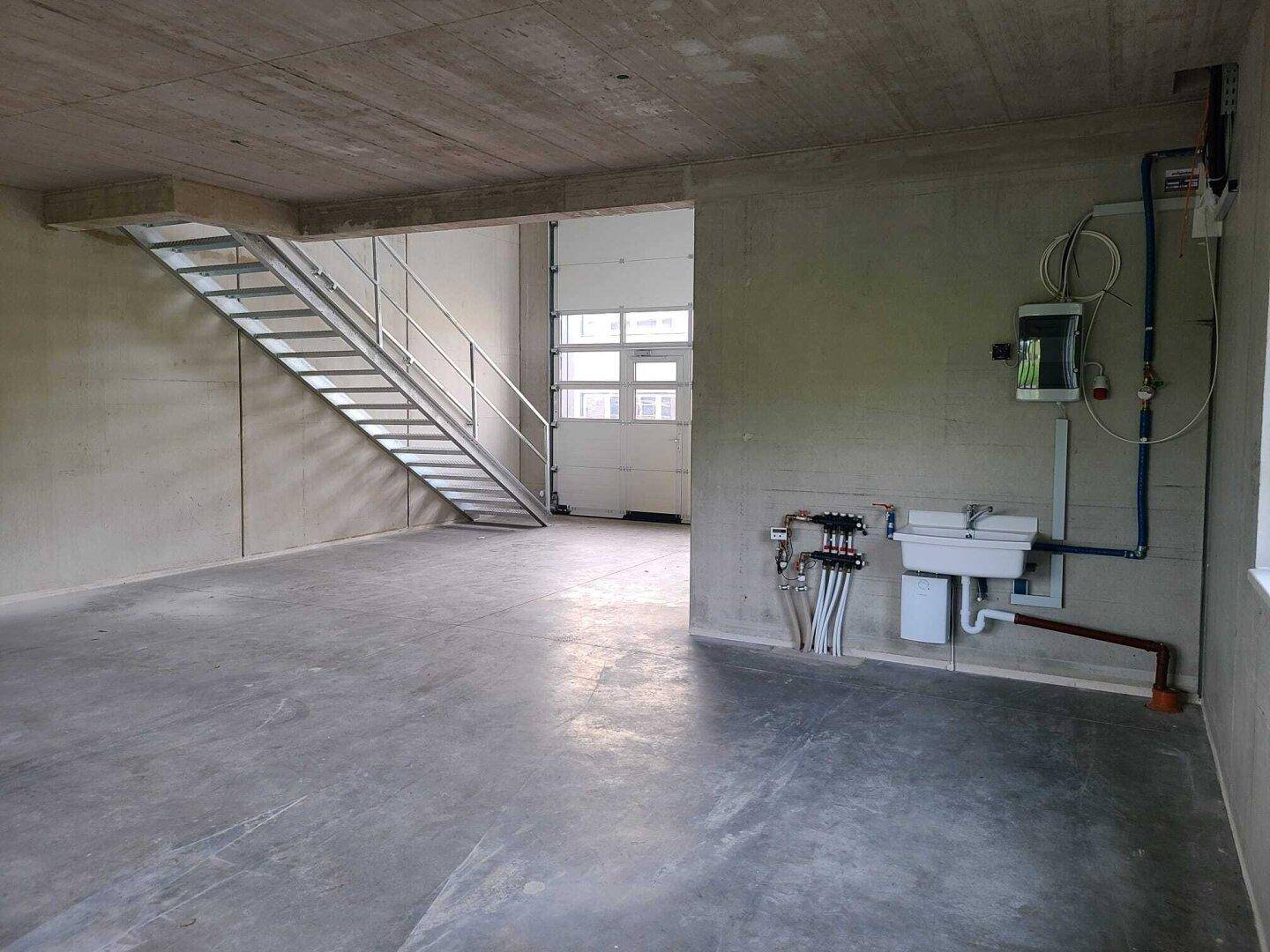 Industrieller Innenraum mit Betonoberflächen, einer Metalltreppe und sichtbaren Installationen an der Wand.