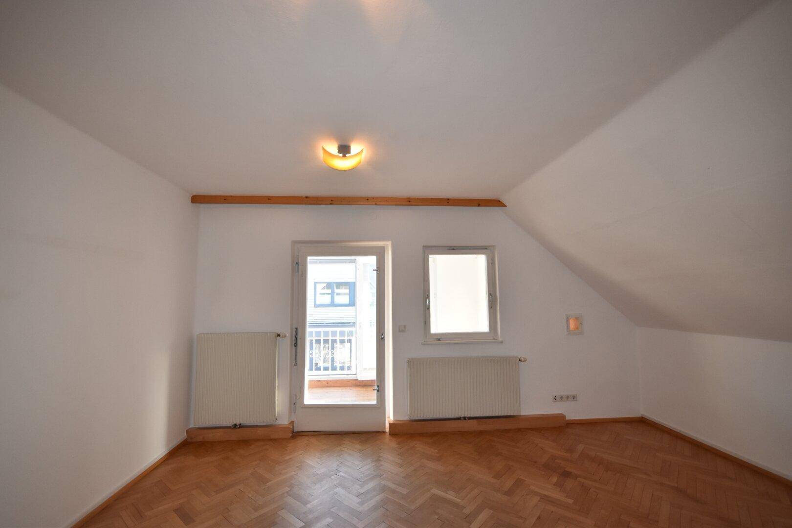Helles Dachzimmer mit Fischgrätparkett, schrägen Wänden und Zugang zu einem kleinen Balkon.