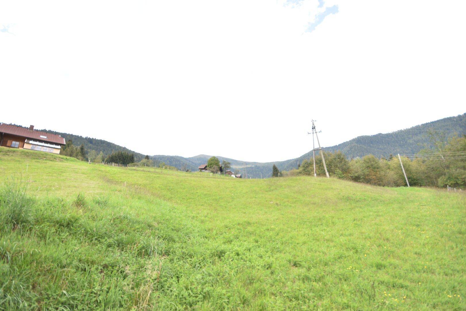 Malerische Landschaft mit grünen Wiesen, einem traditionellen Haus auf dem Hügel und bewaldeten Bergen im Hintergrund.