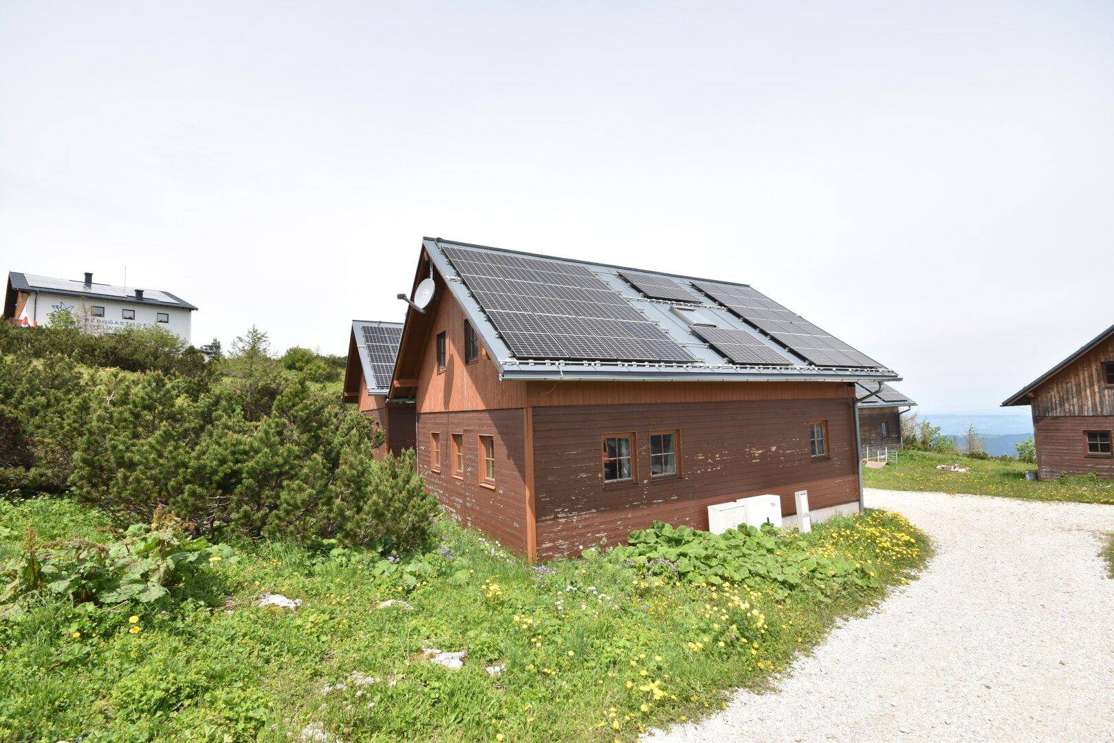 Chalet mit Photovoltaikmodulen auf dem Dach, umgeben von alpiner Vegetation.
