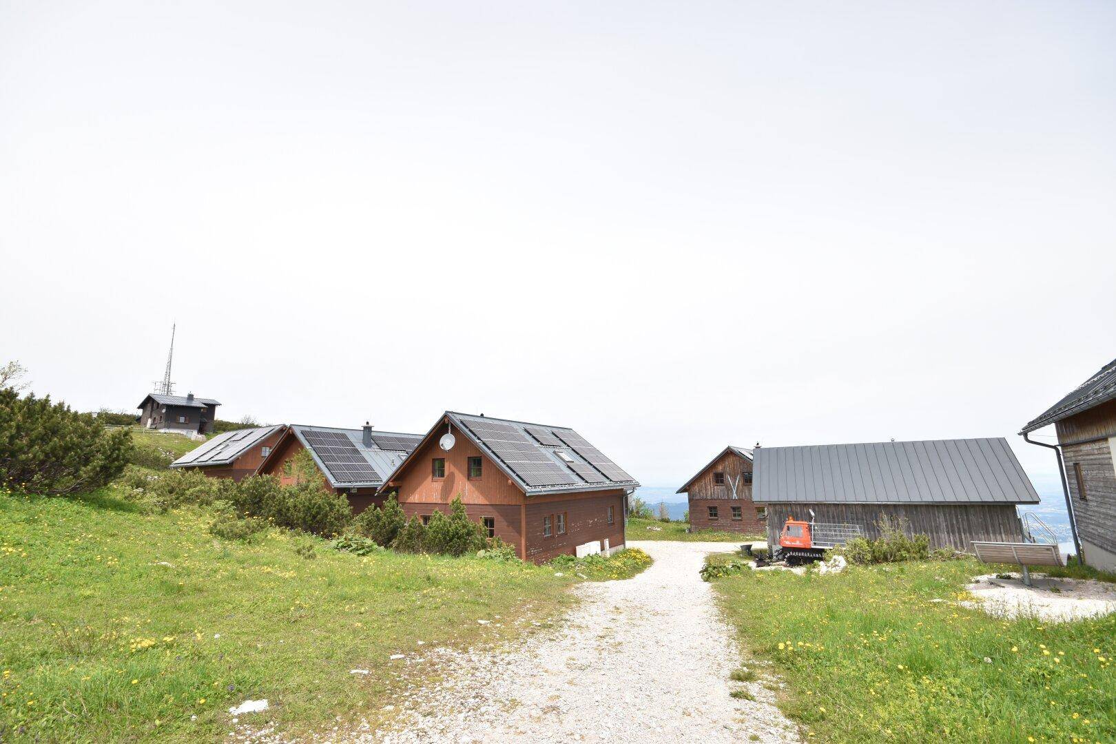 Mehrere Holzhäuser mit Solaranlagen in einer weiten, grünen Berglandschaft.