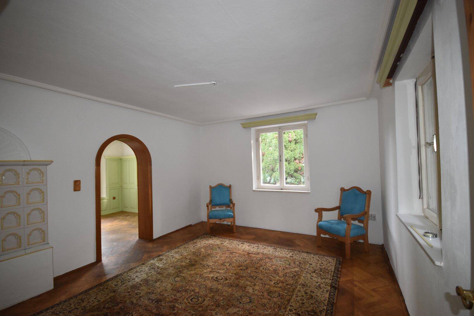 Gemütliches Wohnzimmer mit traditionellem Kachelofen, Parkettboden und zwei blauen Sesseln.