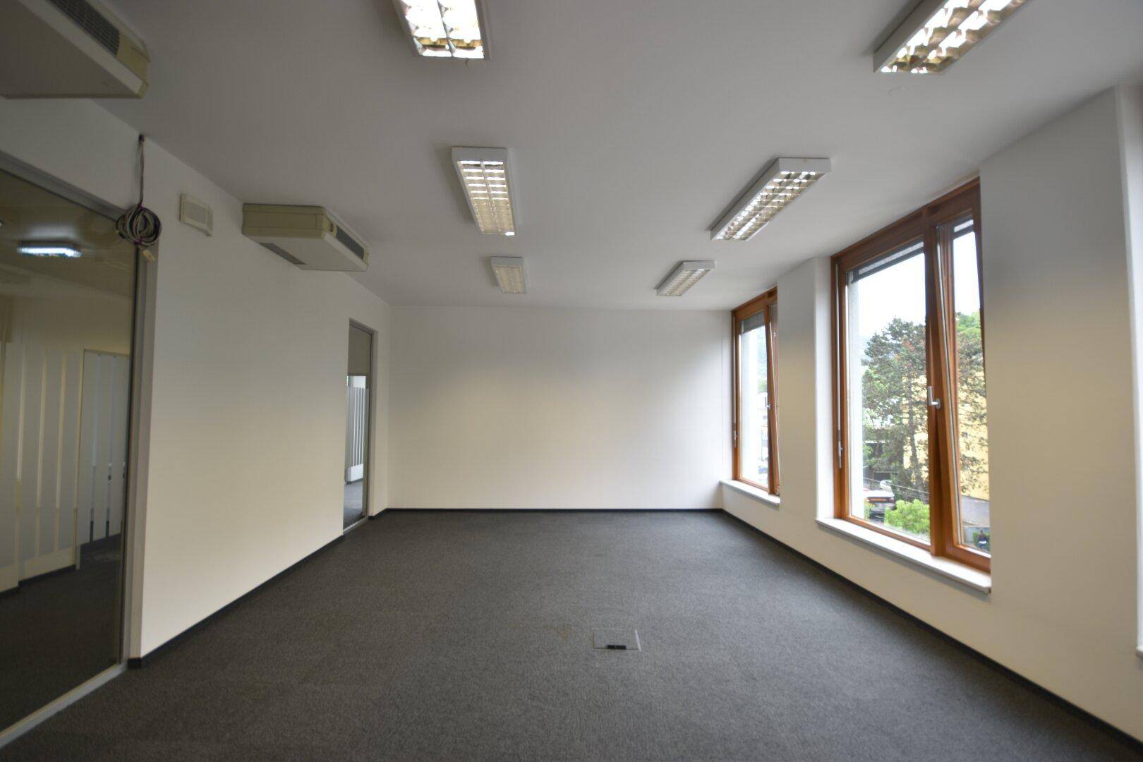 Helles Büro mit Teppichboden, Holzfenstern, Klimaanlage und Blick auf die Umgebung.