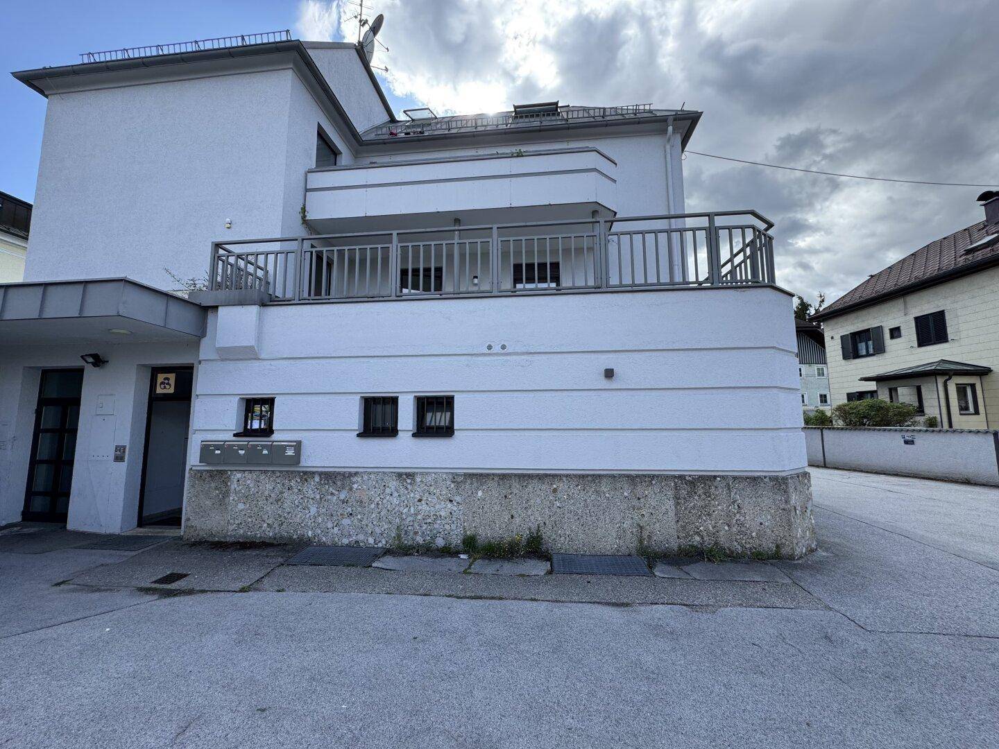 Seitenansicht des weißen Gebäudes mit einem Balkon und grauen Geländern sowie Briefkästen an der Wand.