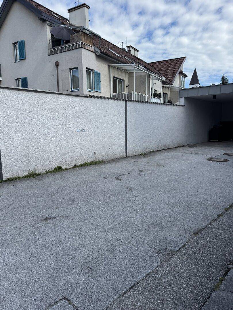 Blick auf eine weiße Mauer mit einem dahinterliegenden Gebäude, das Balkone und eine Garageneinfahrt zeigt.