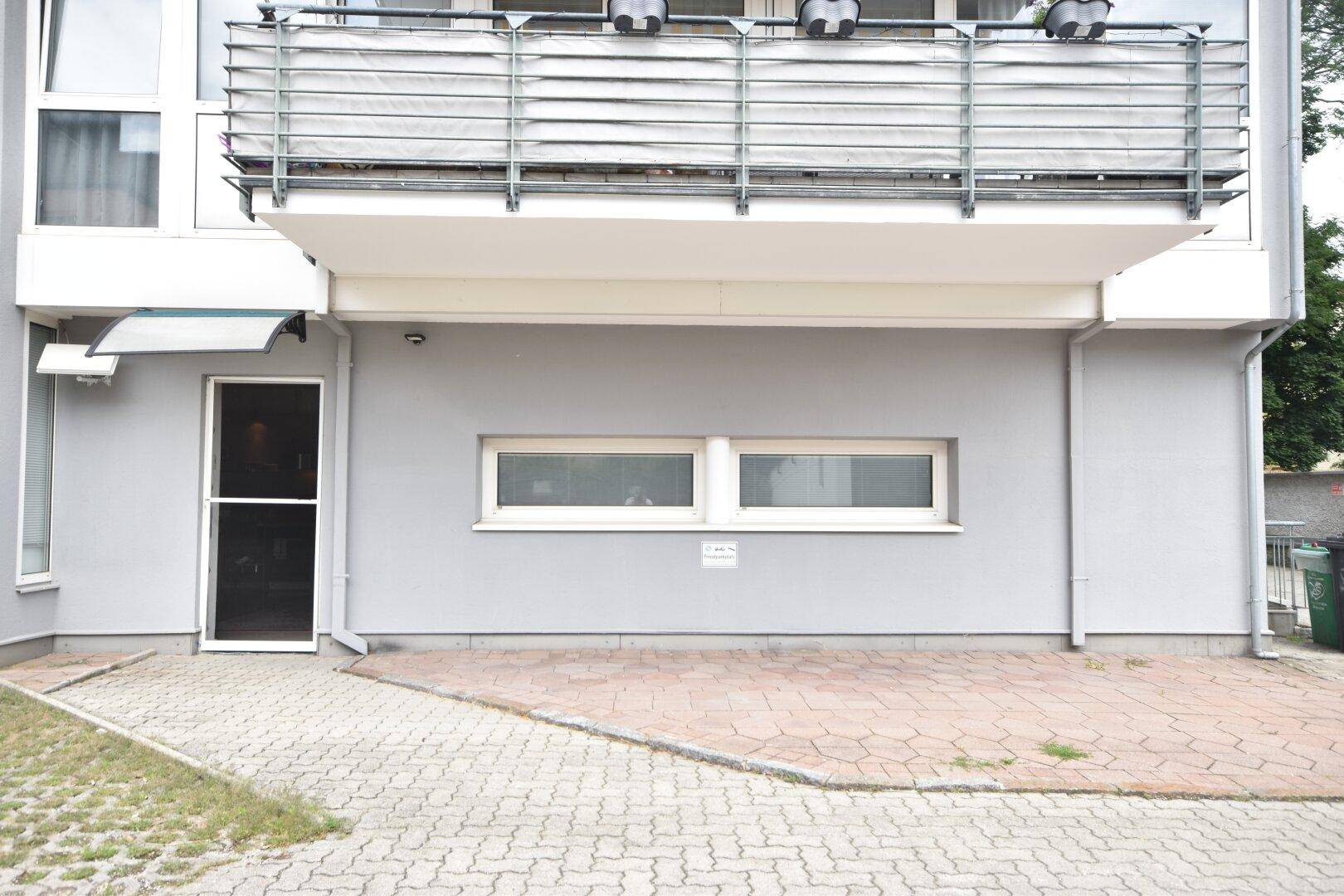 Gebäudeeingang mit gepflastertem Vorplatz und kleinen Fenstern im Untergeschoss.