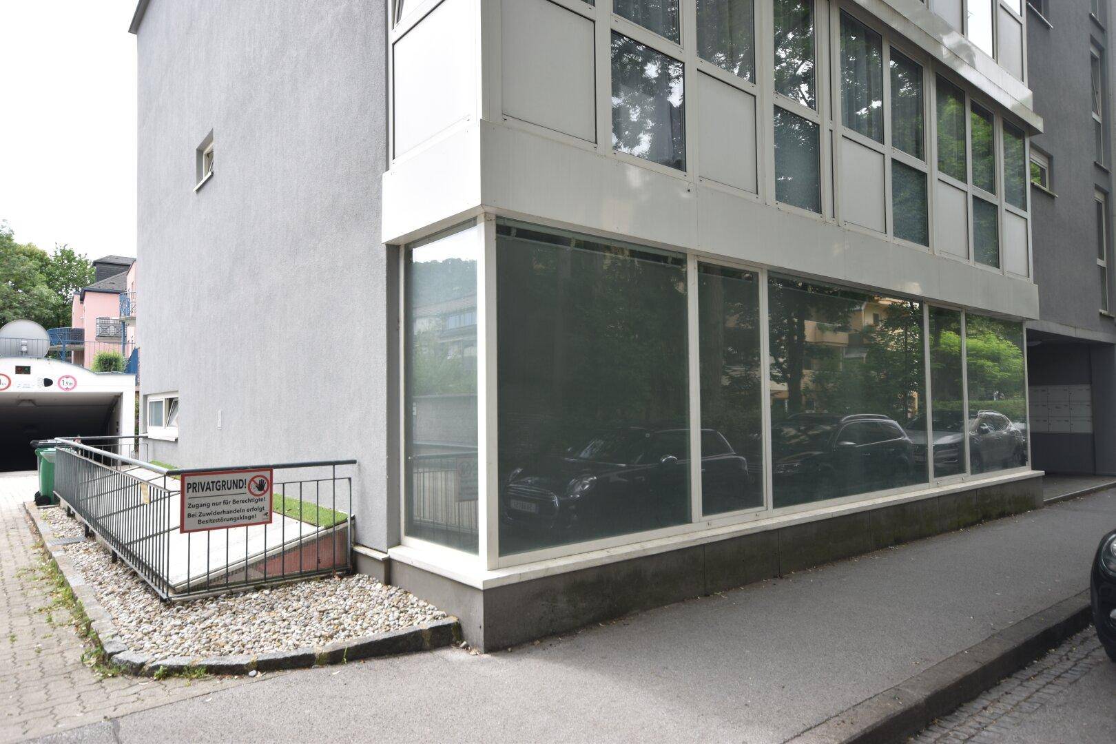 Gebäudeecke mit großen Schaufenstern im Erdgeschoss und einem Zugang zum Untergeschoss mit Rampe.