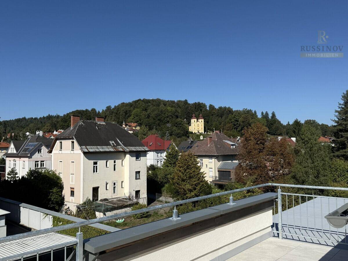 Blick von einer Terrasse auf Wohngebäude, grüne Hügel und eine historische Kirche.