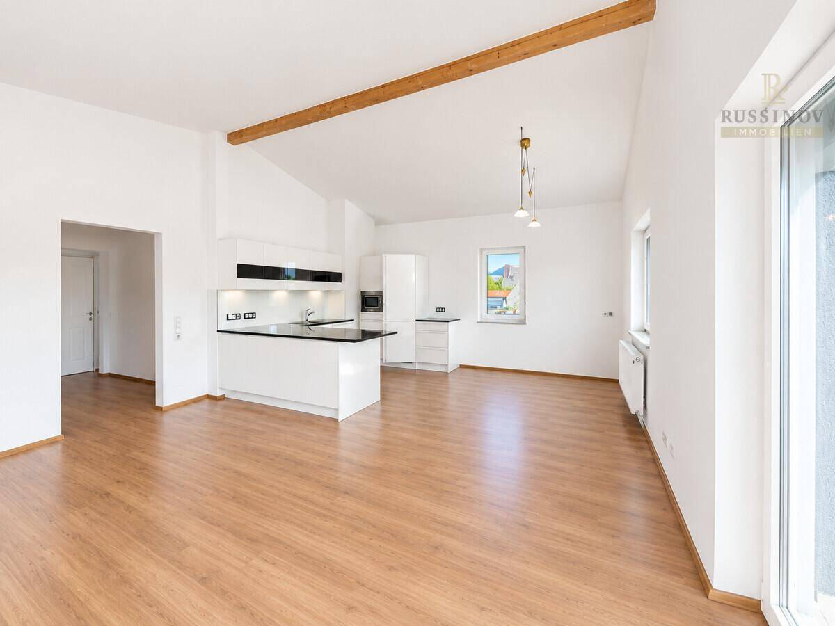 Geräumiger Wohnbereich mit integrierter Küche, Laminatboden und sichtbarem Holzbalken unter der Decke.