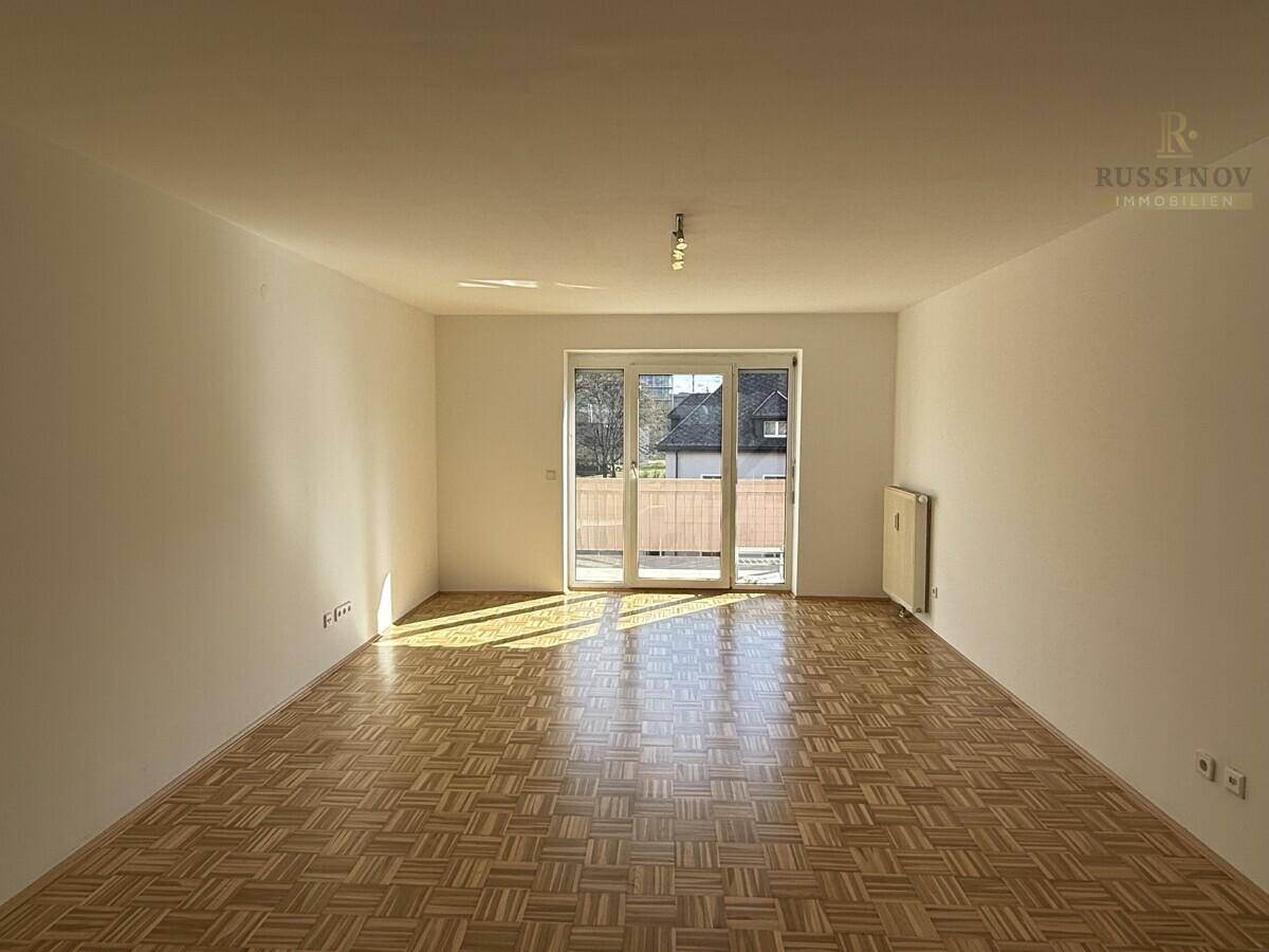 Helles Wohnzimmer mit Parkettboden und Zugang zu einem Balkon, sonnendurchflutet.