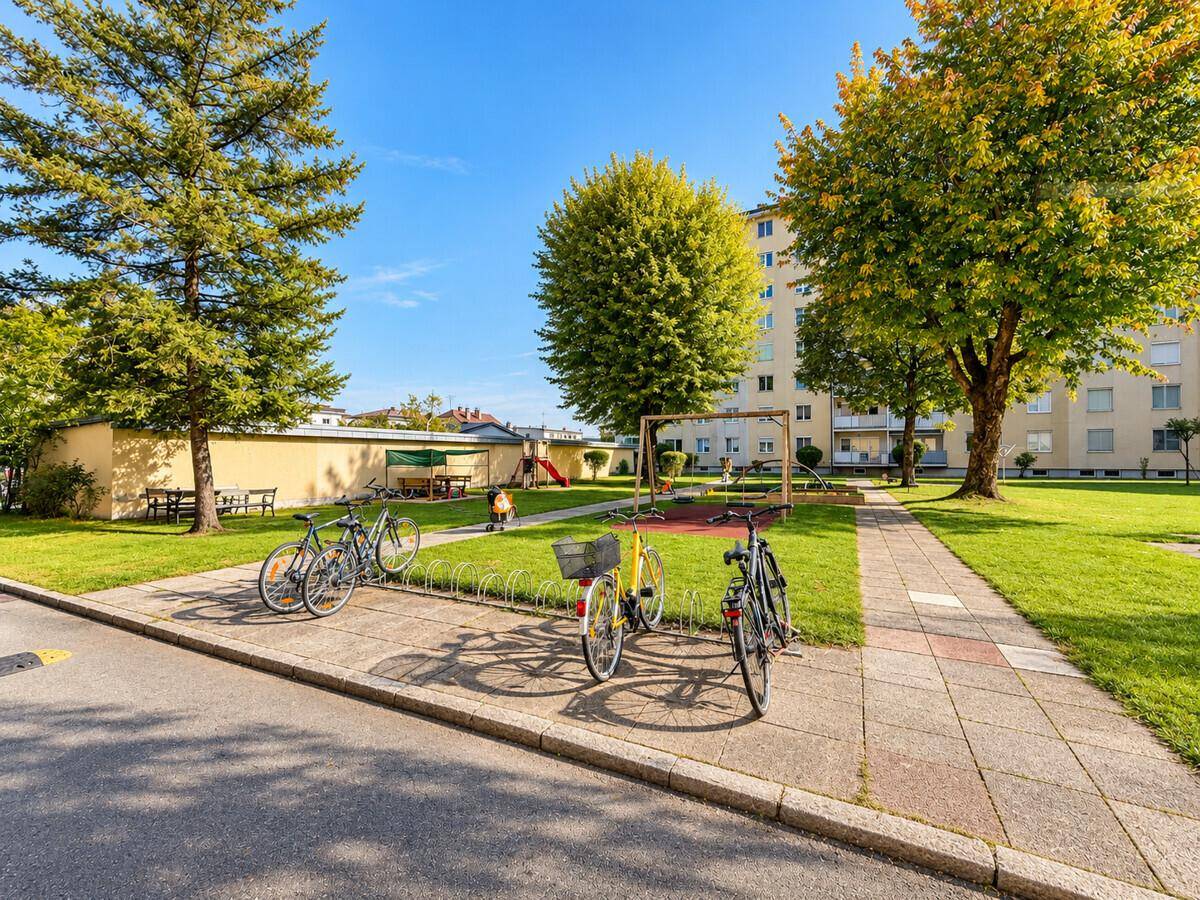 Grüne Gemeinschaftsfläche mit Spielplatz, Bäumen und Fahrradständern vor einem Wohngebäude.