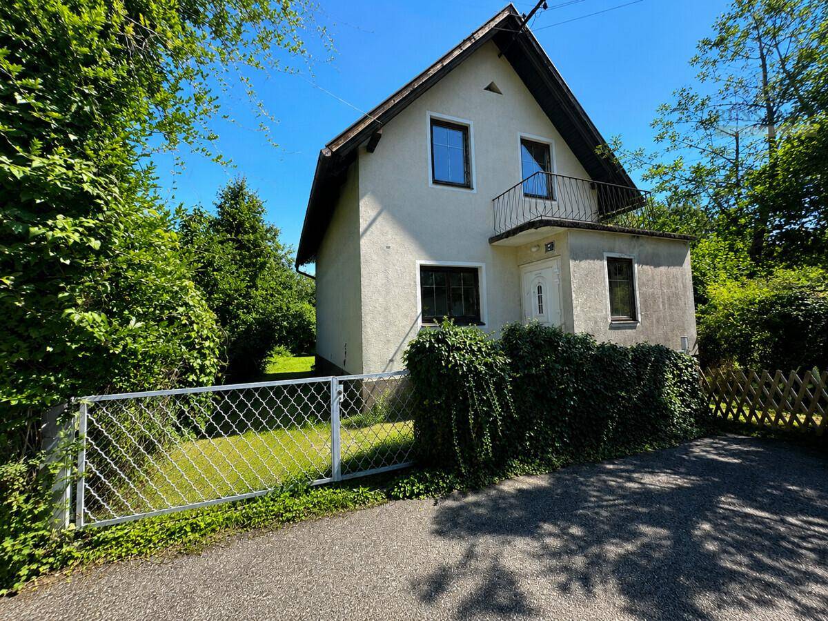Vorderansicht eines Hauses mit kleinem Balkon, umgeben von viel Grün und einem Zaun.