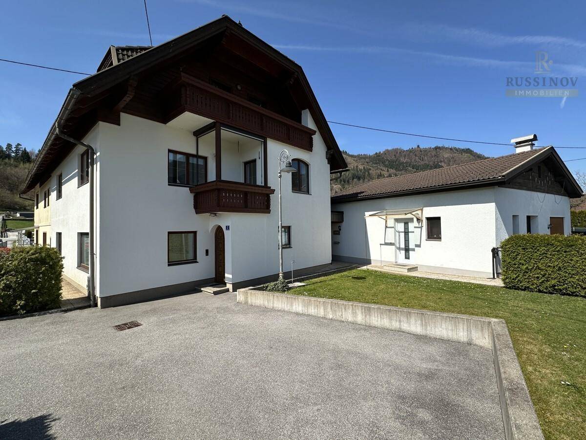 Traditionelles Haus mit Holzbalkon und großem Vorplatz, umgeben von grüner Natur.