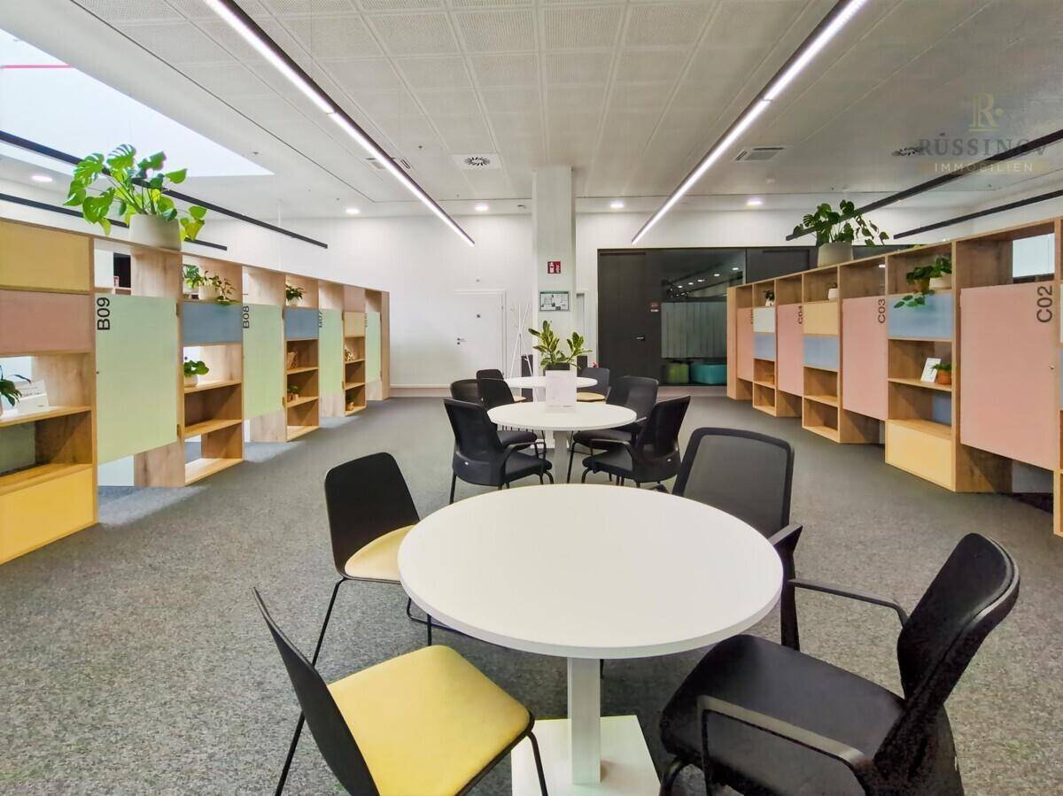 Moderner Kollaborationsbereich in einem Büro mit runden Tischen und Stühlen, umgeben von farbigen Regalen.
