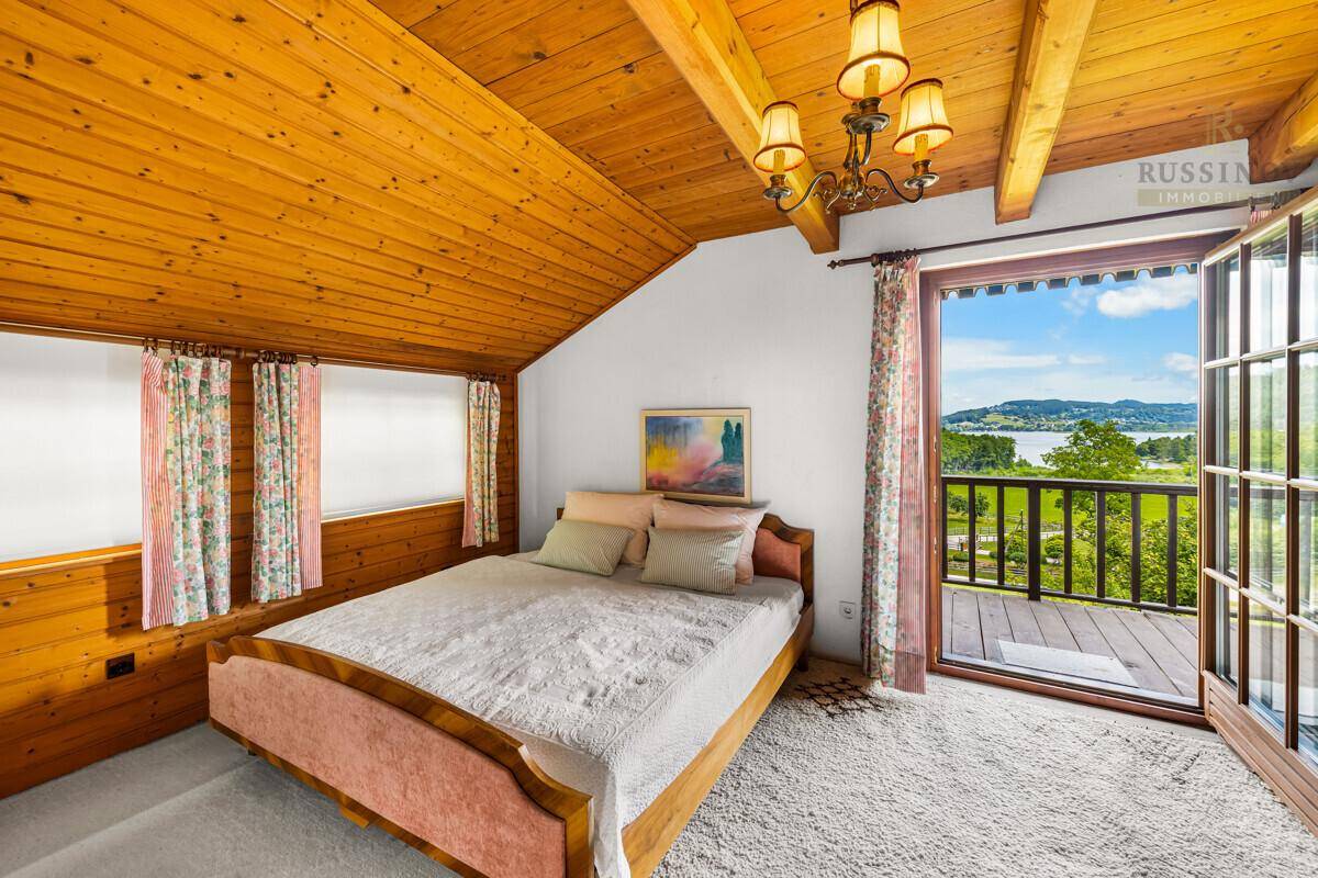 Gemütliches Schlafzimmer mit Holzbalkendecke, Teppichboden und Zugang zu einem Balkon mit Seeblick.