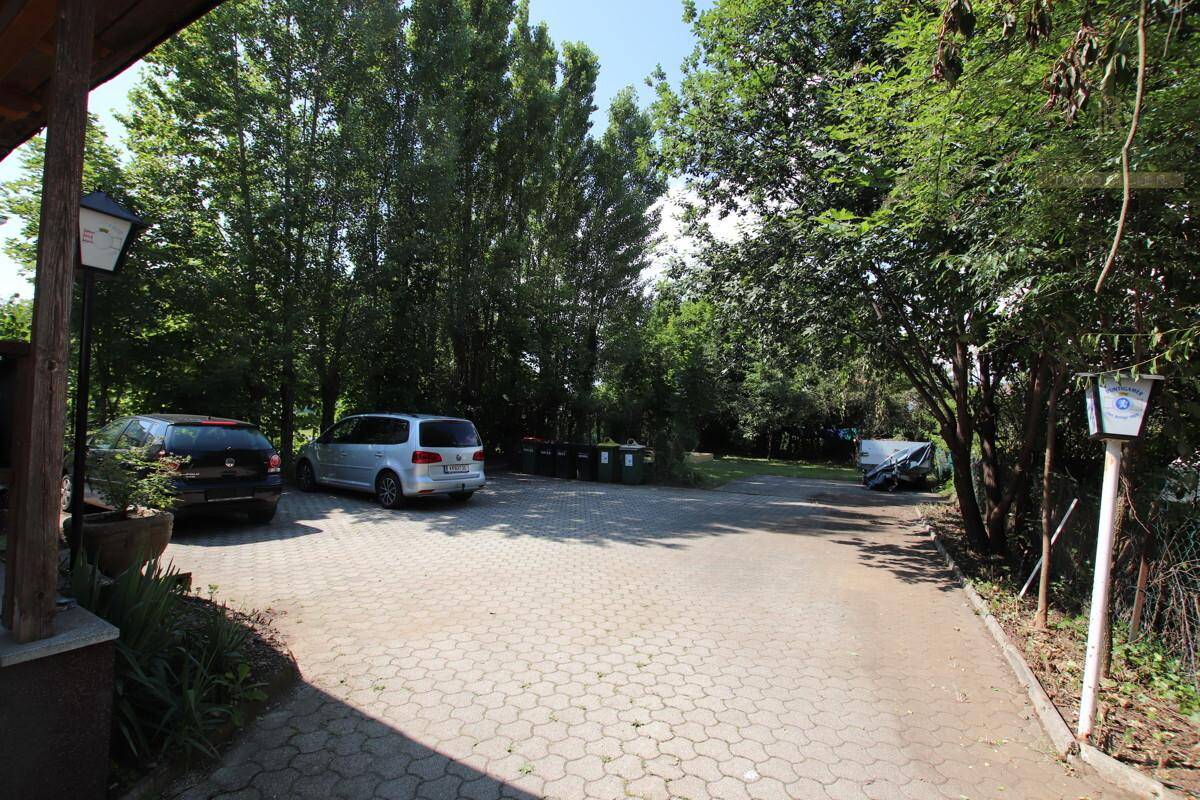 Großer gepflasterter Parkplatz mit mehreren abgestellten Fahrzeugen, umgeben von hohen Bäumen.