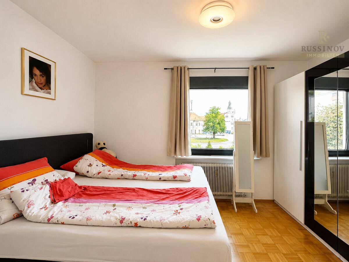 Schlafzimmer mit Doppelbett, Parkettboden und Fenster mit Blick ins Grüne und auf ein Gebäude.
