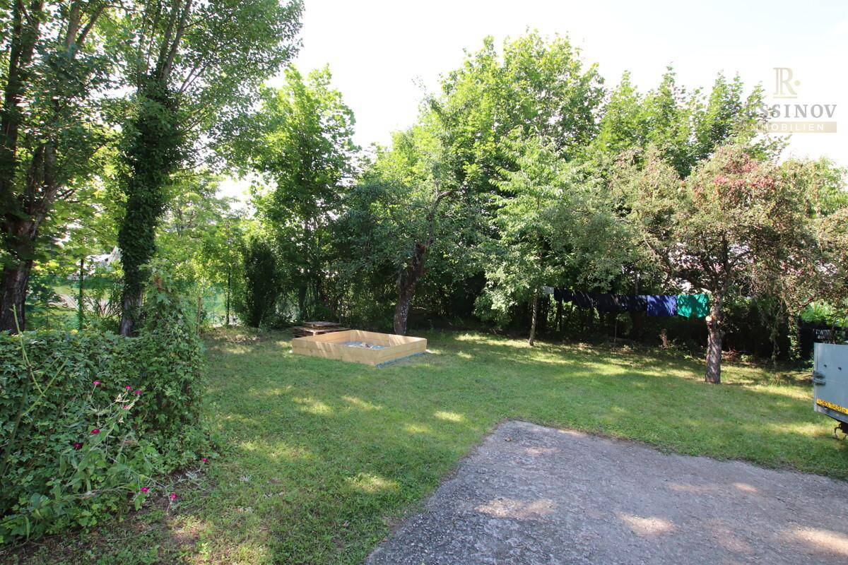 Sonniger Garten mit Rasen, Bäumen und einem Sandkasten, im Hintergrund Wäsche auf einer Leine.