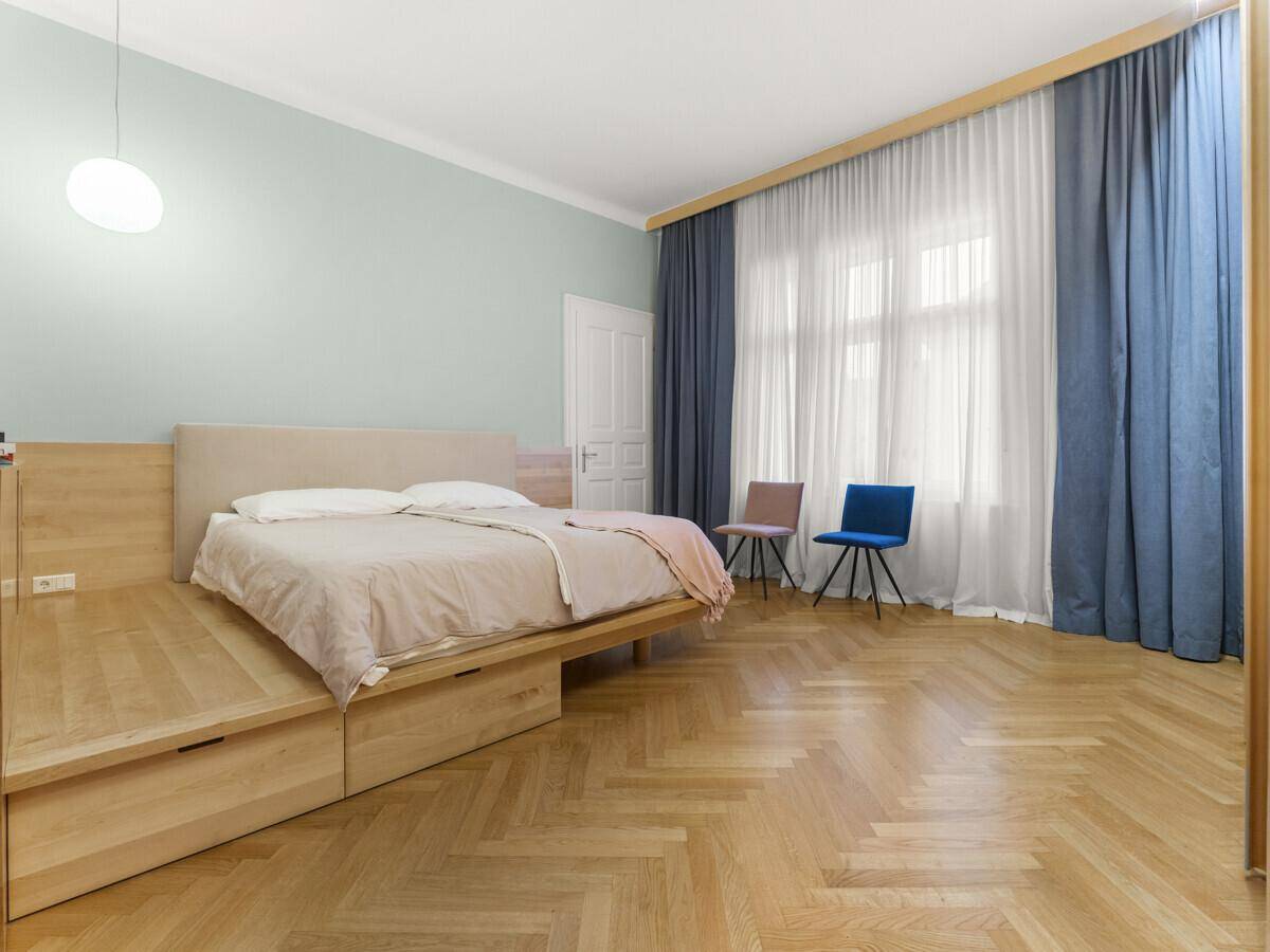 Helles Schlafzimmer mit Holzbett, Fischgrätparkett und großen Fenstern mit blauen Vorhängen.