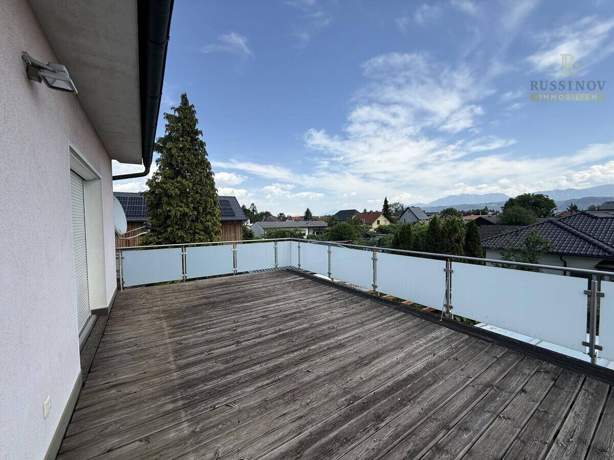 Großer Balkon mit Holzboden und Glasgeländer, bietet einen weiten Blick auf die umliegende Landschaft und Berge.