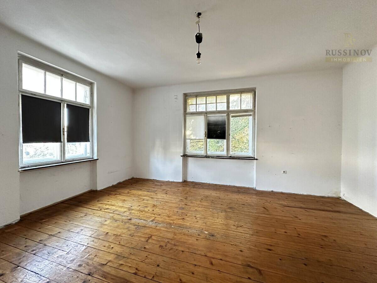 Geräumiges, leeres Zimmer mit Holzdielenboden und großen Fenstern, bereit zur Renovierung.