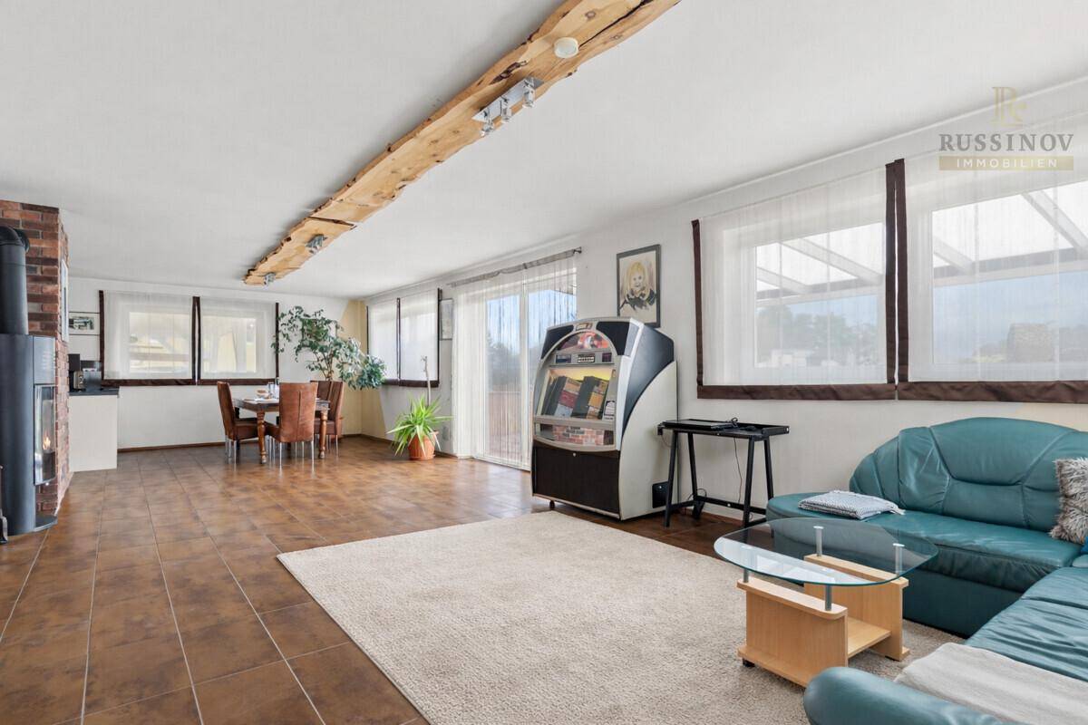 Geräumiger Wohn-Essbereich mit Holzbalken, Kamin und großen Fenstern, die viel Licht hereinlassen.