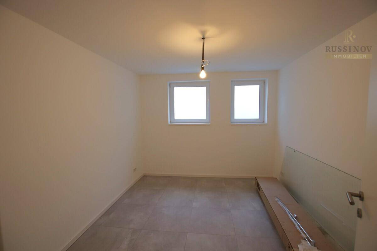 Heller, unmöblierter Raum mit zwei Fenstern, Fliesenboden und einer Glasbrüstung.