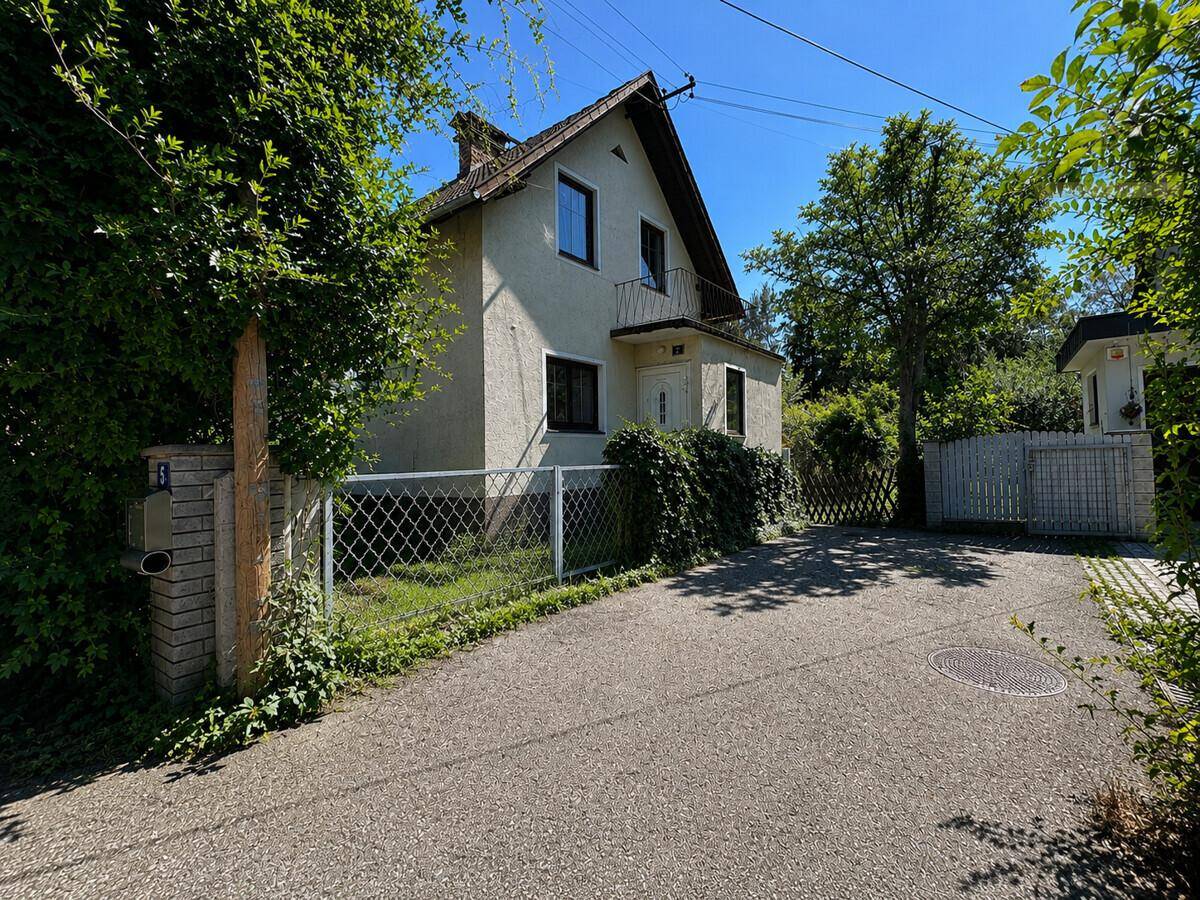 Hausansicht von der Einfahrt aus, umgeben von Bäumen und einem Zaun.