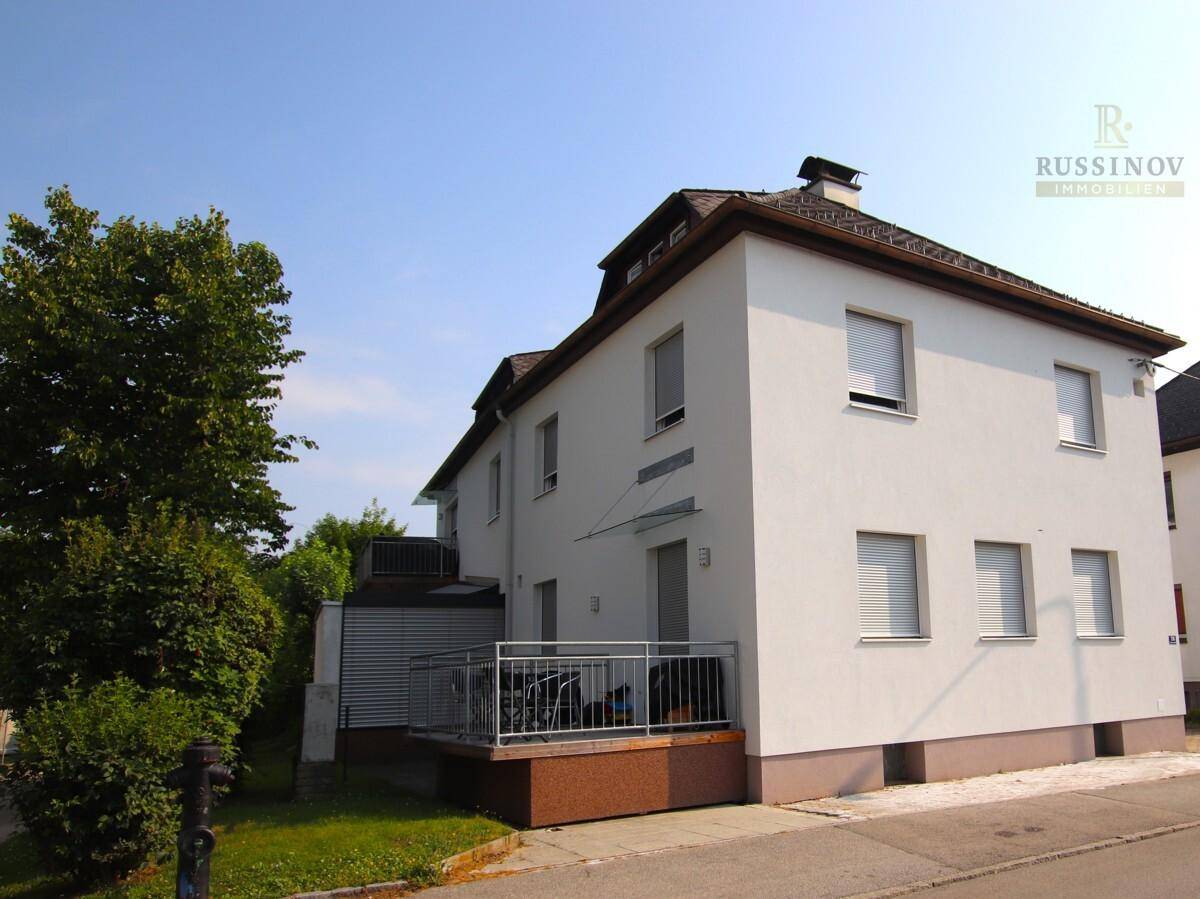 Seitenansicht eines weißen Gebäudes mit Balkon und Rollläden, umgeben von Bäumen und einer Straße.