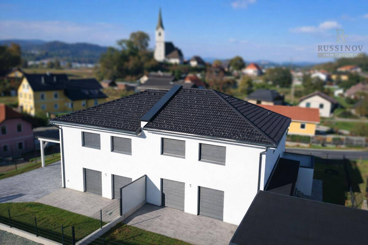 Luftaufnahme eines modernen Doppelhauses mit dunklem Dach und Blick auf die umliegende Landschaft und Kirche.