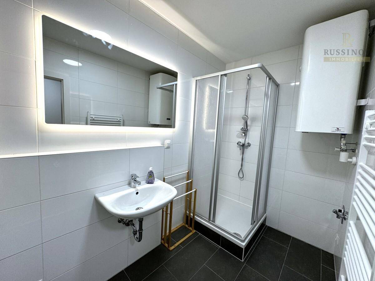 Modernes Badezimmer mit Duschkabine, beleuchtetem Spiegel und weißen Fliesen.