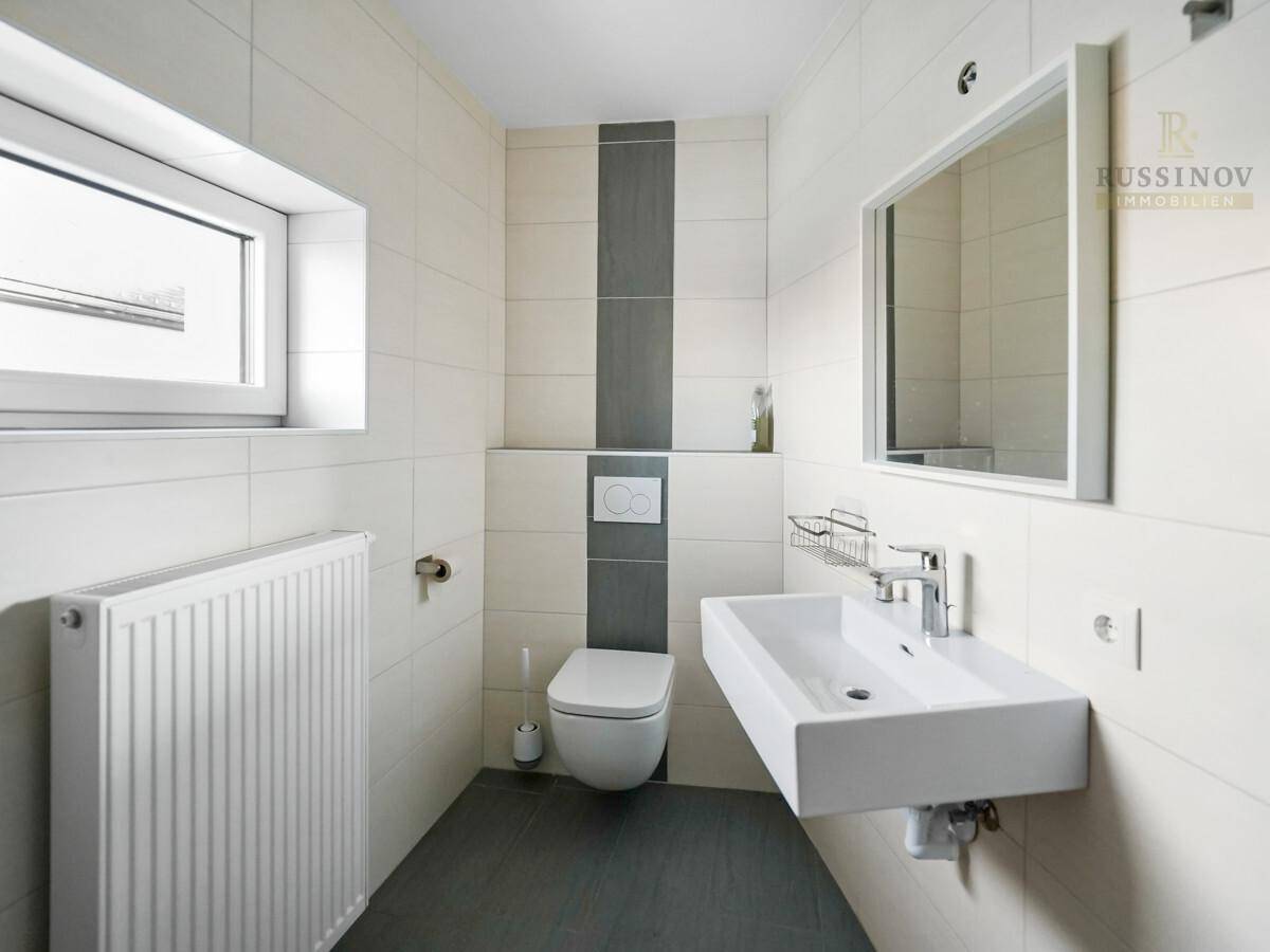 Helles Badezimmer mit modernem Wand-WC, Waschbecken und Fenster, gefliest in Weiß und Grau.