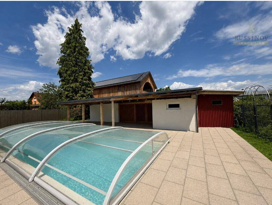 Außenbereich mit überdachtem Pool, gepflasterter Fläche und einem Carport, umgeben von viel Grün.