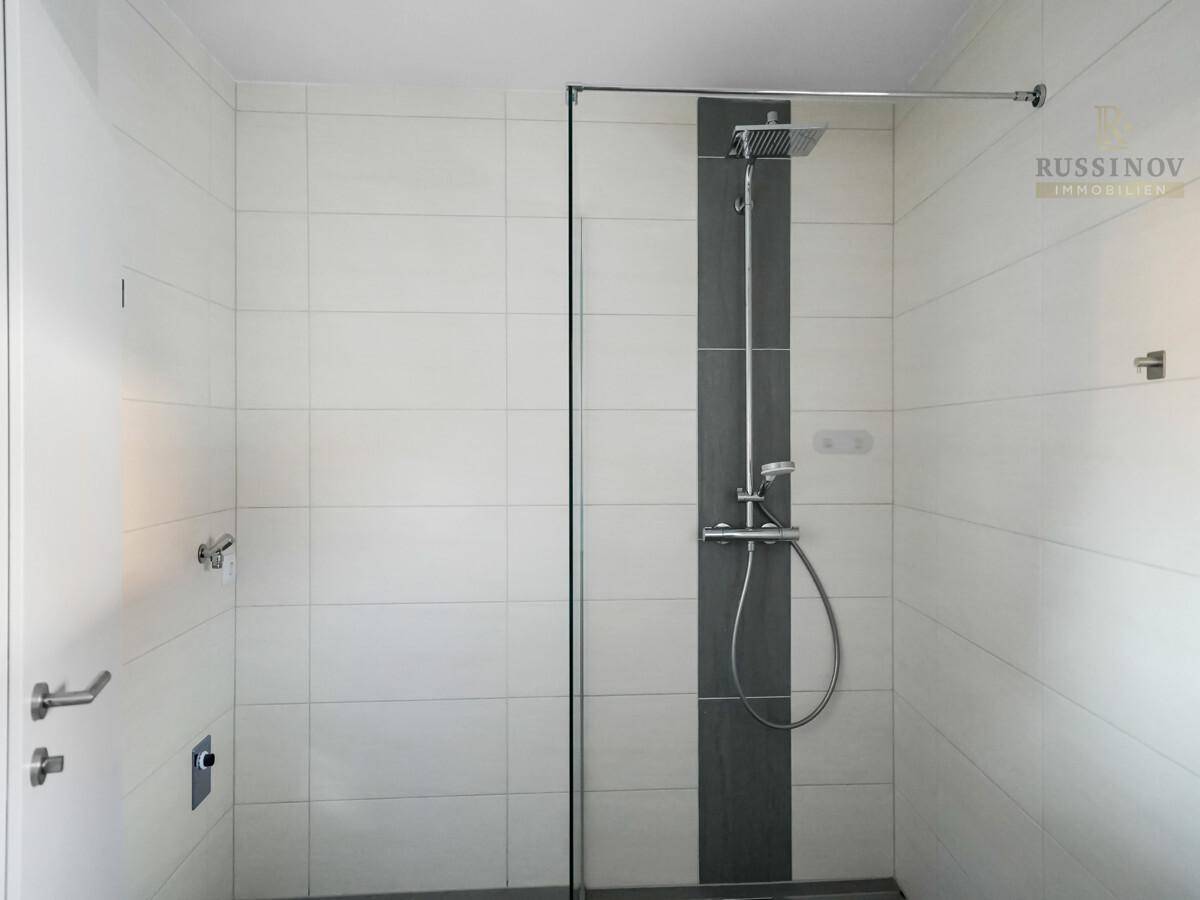 Moderne Duschkabine mit Glaswand und Regenduschkopf, gefliest in Weiß und Grau.