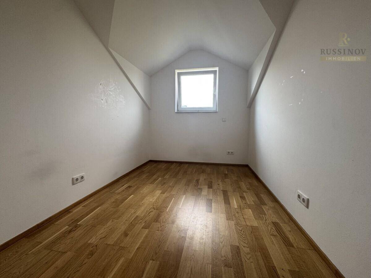 Kleineres Zimmer im Dachgeschoss mit Holzboden, weiß gestrichenen Wänden und einem Fenster.