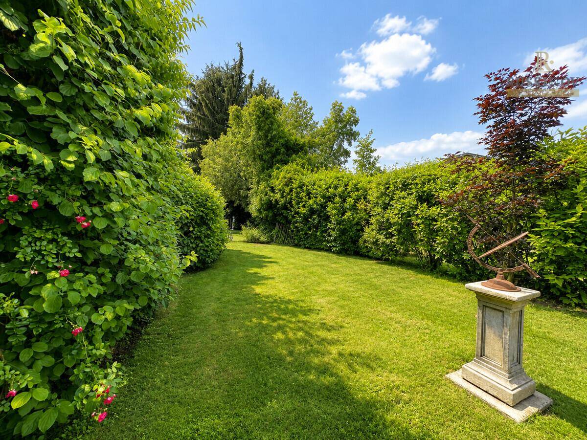 Gepflegter Gartenweg zwischen hohen Hecken und Bäumen mit einer Sonnenuhr als Dekoration.