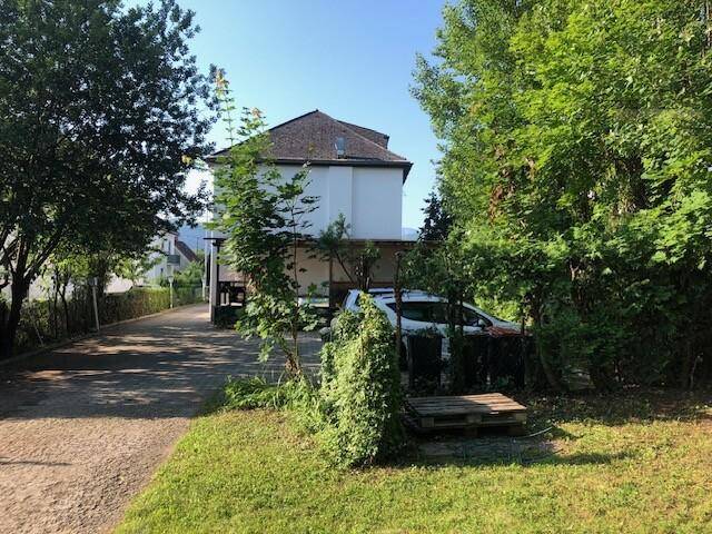 Zufahrt zum Gebäude mit einem geparkten Auto, umgeben von üppigem Grün und hohen Bäumen.