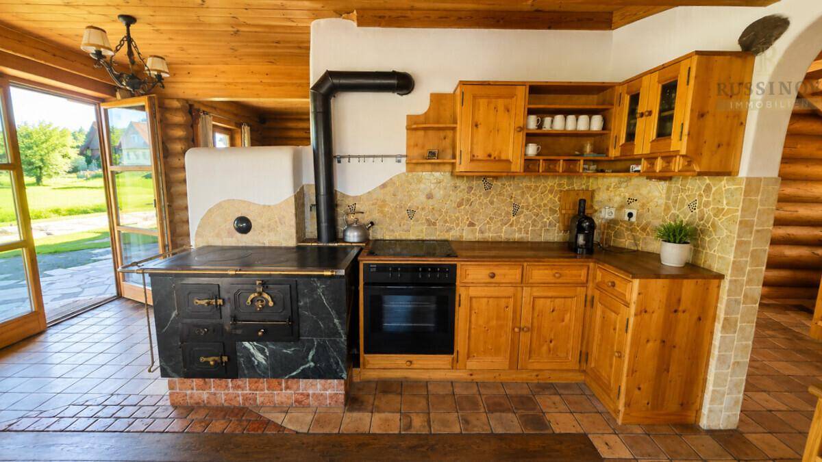 Rustikale Holzküche mit traditionellem Kachelofen und Fliesenboden, offen zum Gartenbereich.