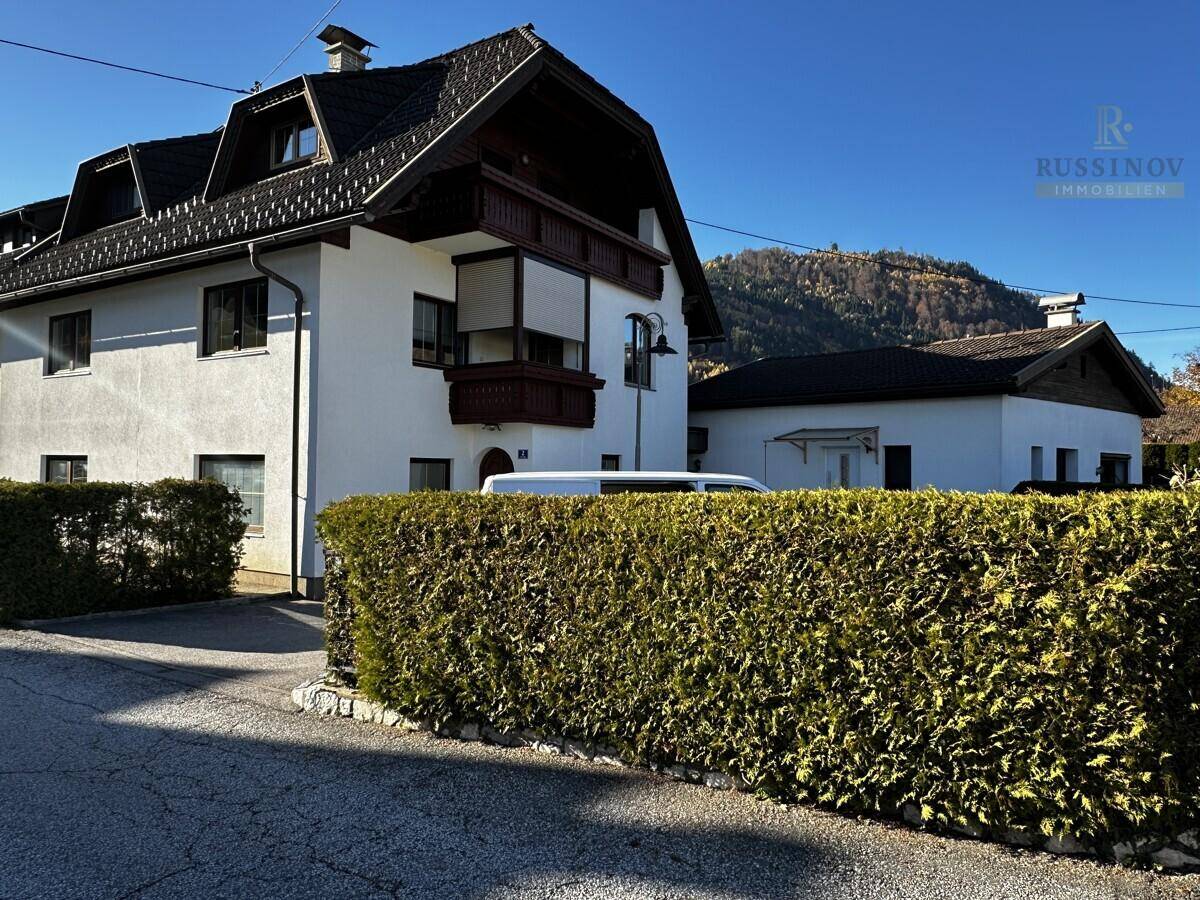 Ansicht des Hauses mit traditioneller Architektur, Holzbalkonen und gepflegter Hecke vor Bergkulisse.