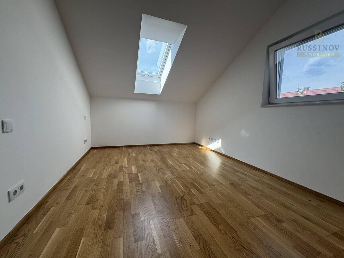 Geräumiges Zimmer im Dachgeschoss mit Holzboden, schrägen Wänden und viel Tageslicht durch Fenster und Dachluke.