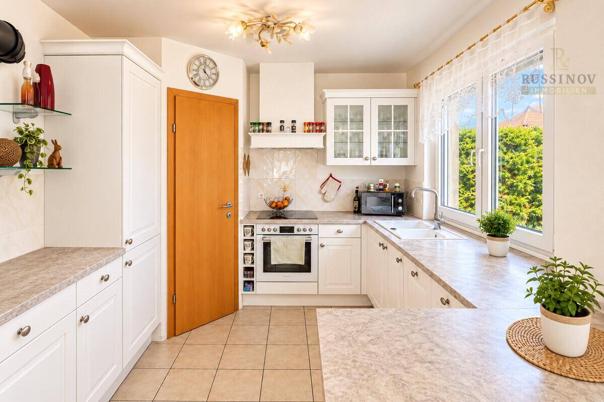 Funktionale weiße Küche mit Fliesenboden, Backofen und Fenster mit Blick ins Grüne.