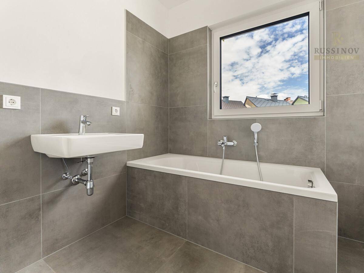 Modernes Badezimmer mit grauen Fliesen, Badewanne, Wandwaschbecken und Fenster.