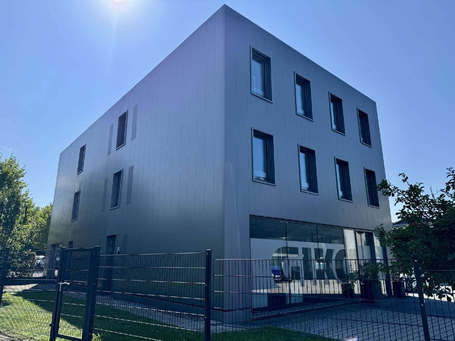 Modernes Bürogebäude mit grauer Fassade, großen Fenstern und einem Zaun im Vordergrund.