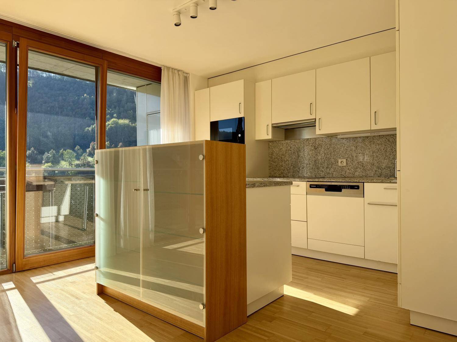 Helle Küche mit Granitarbeitsplatte und einem freistehenden Schrank, viel Sonnenlicht durch große Fenster.