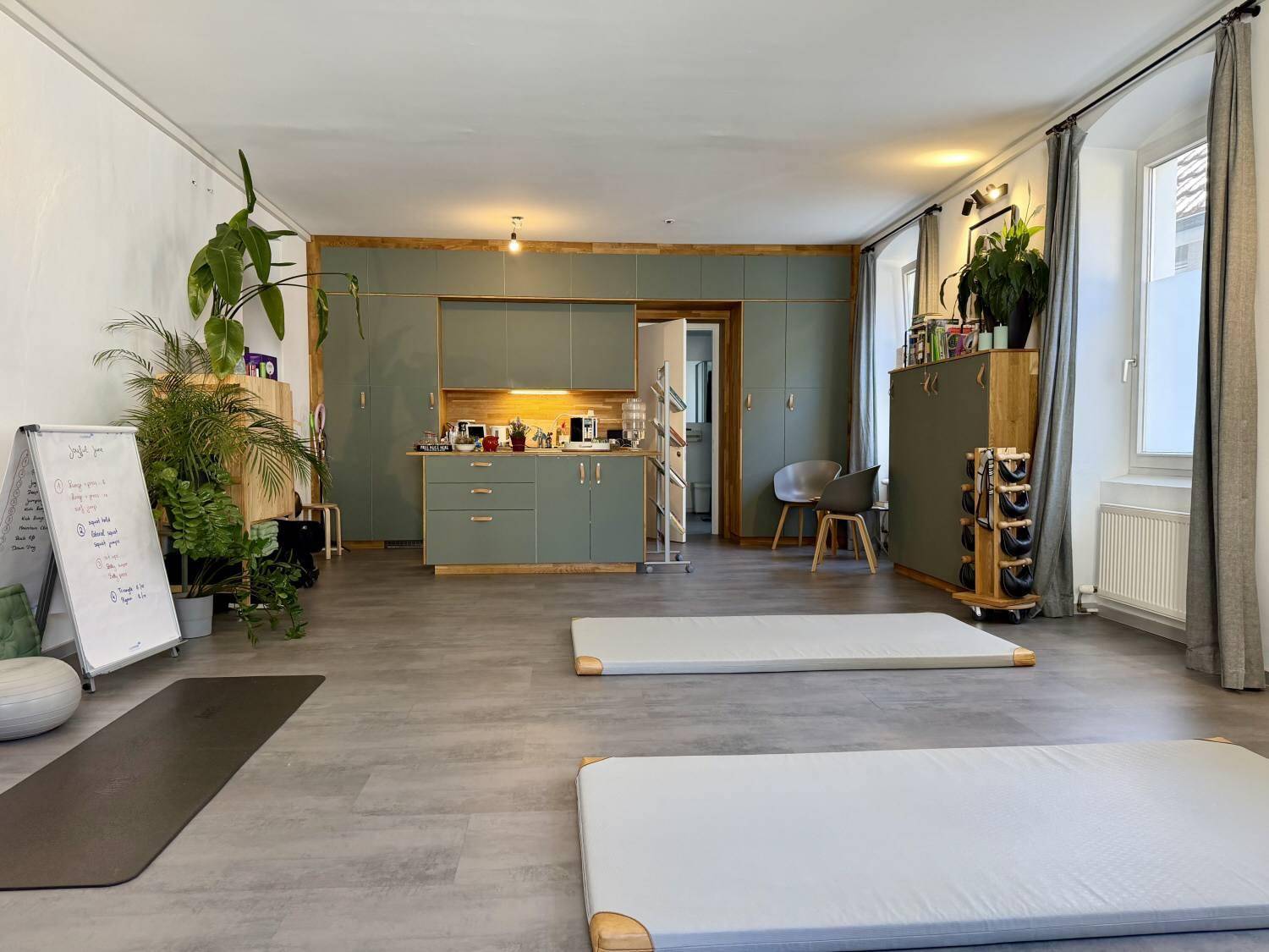 Geräumiger offener Studiobereich mit integrierter moderner Küchenzeile, Trainingsmatten und vielen Pflanzen für eine angenehme Atmosphäre.