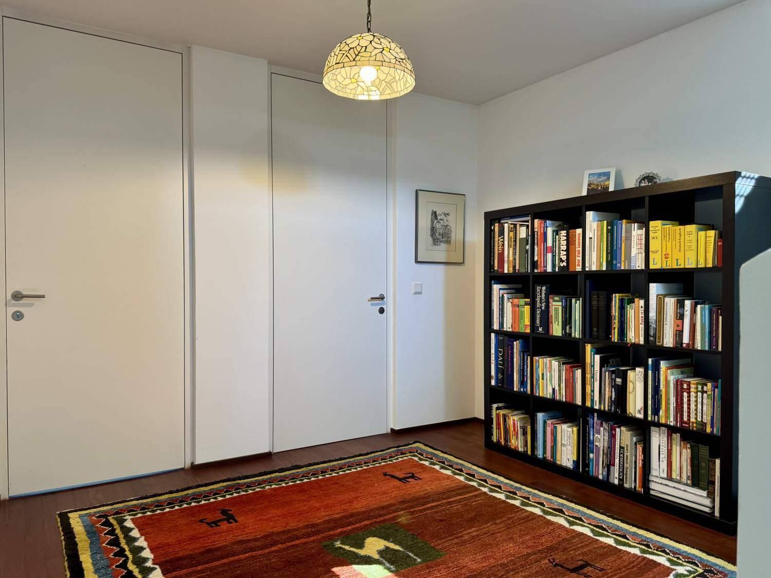 Zimmer mit Bücherregal, Holzboden und farbenfrohem Teppich, ideal als Büro oder Gästezimmer.