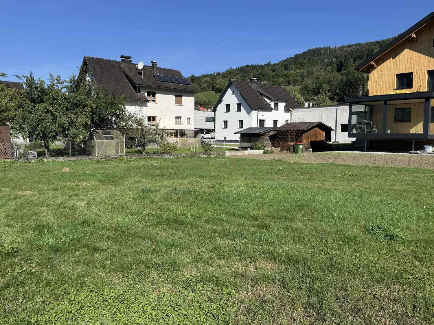 Sonnige Rasenfläche mit neuem Holzhaus und weiteren Wohnhäusern vor bergiger Kulisse.