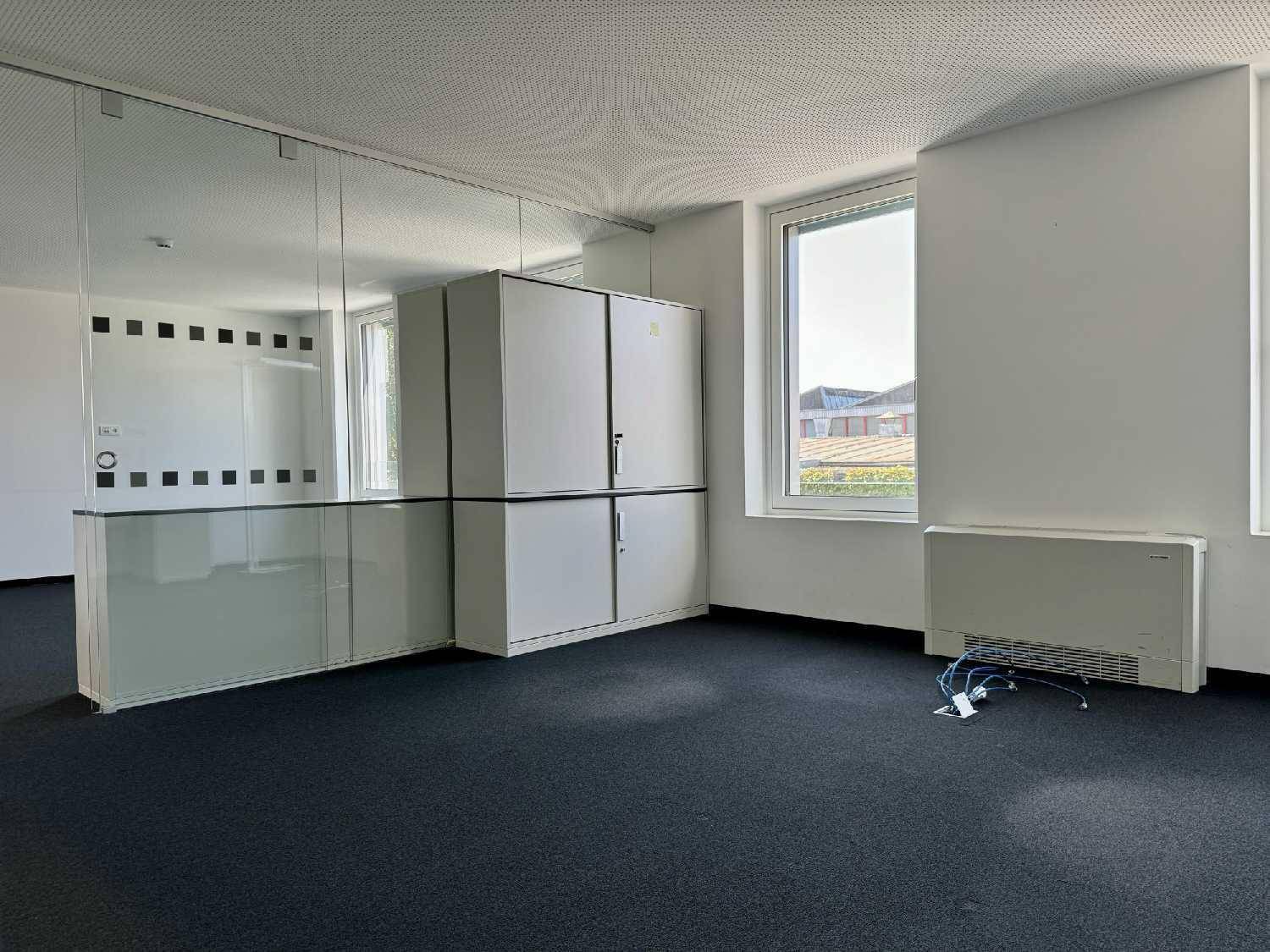 Heller Bürobereich mit großen Fenstern, weißen Schränken und dunklem Teppichboden.