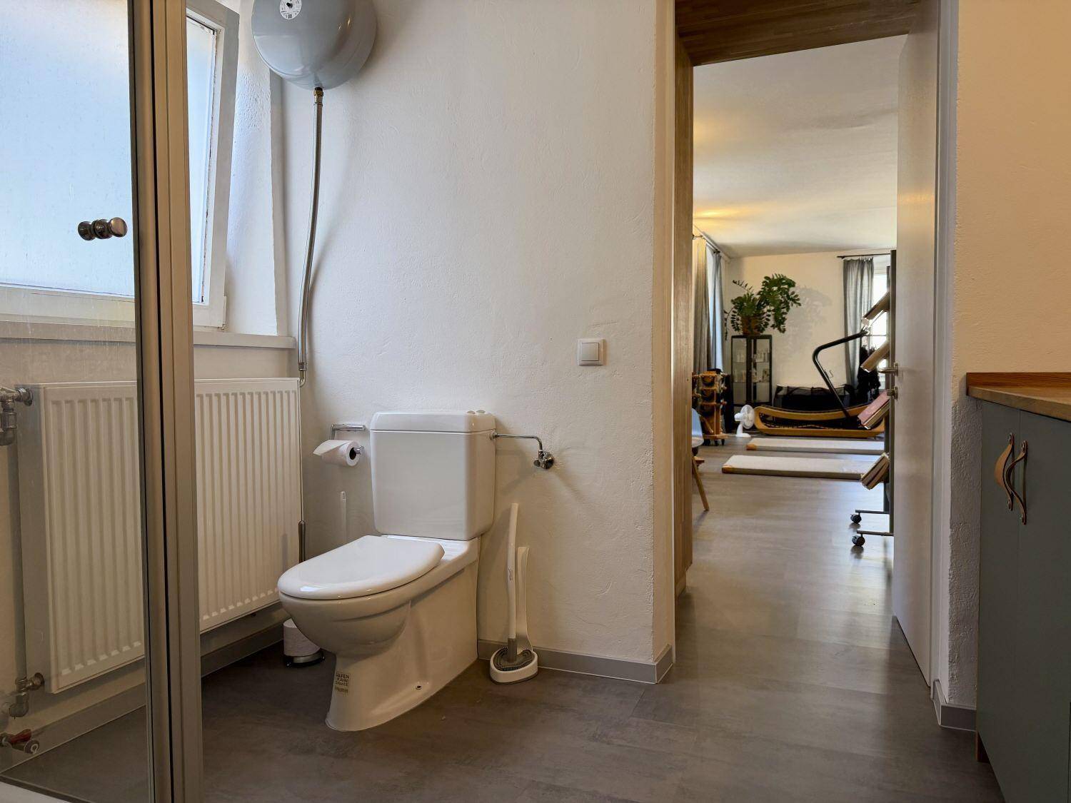 Toilettenbereich im Badezimmer mit Fenster, Heizkörper und einem Blick in den angrenzenden Studiobereich.