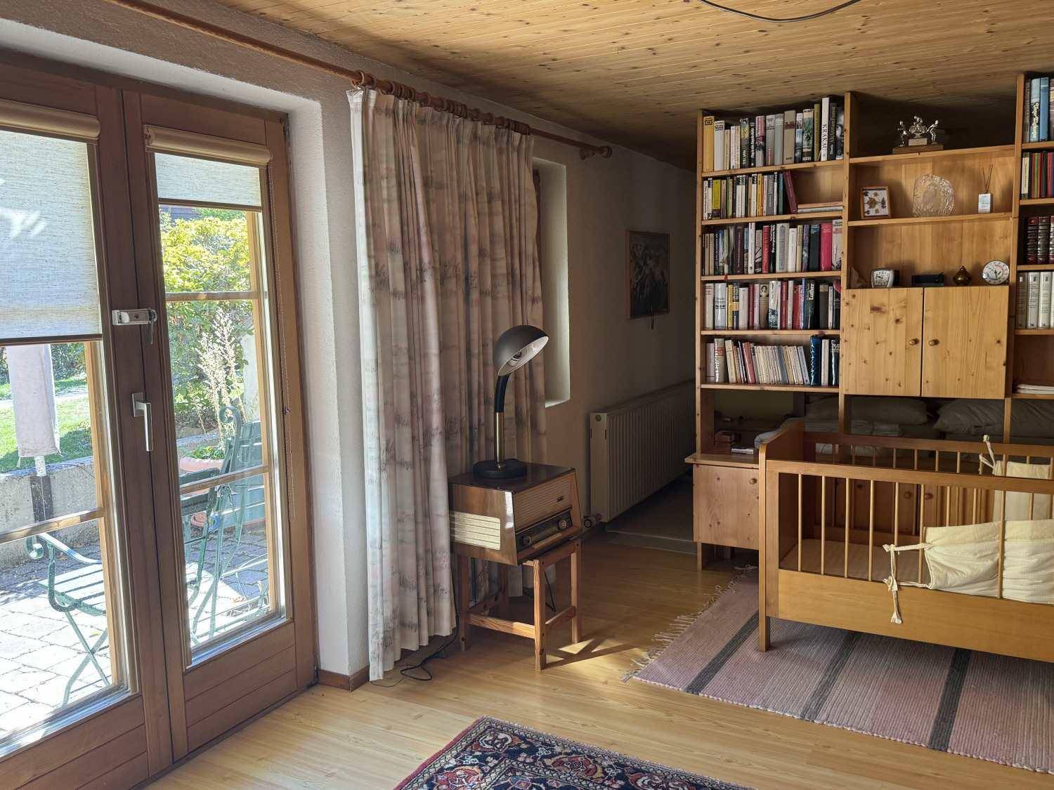 Gemütlicher Wohnbereich mit Holzbalkendecke, großem Bücherregal und Zugang zum Garten.